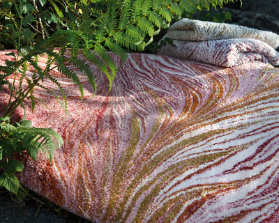 A textured rug with red, pink, and yellow swirls lies on the ground next to a folded matching rug, partially covered by green fern leaves.