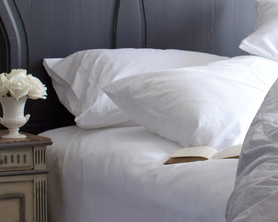 A neatly made bed with white pillows and sheets, an open book on the bed, and a small vase with white flowers on a bedside table.