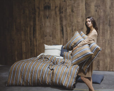 A woman in a brown dress holds two striped pillows near a bed with matching striped bedding and a wooden wall backdrop.