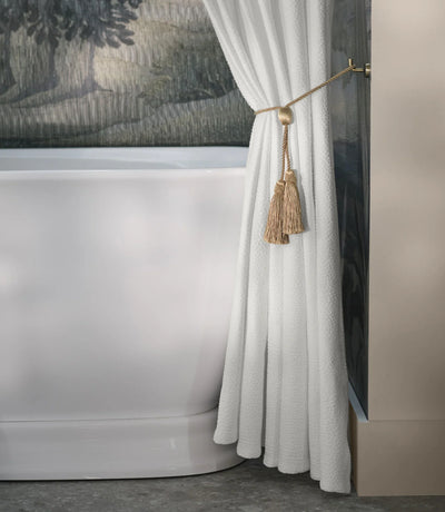 Close-up of a white bathtub partially covered by a white curtain with gold tassel tiebacks, set against a wall with a textured, scenic mural.