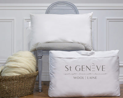 A white pillow on a gray chair, a large pillow bag labeled St GENÈVE WOOL | LAINE, and a basket of wool sit against a white paneled wall.