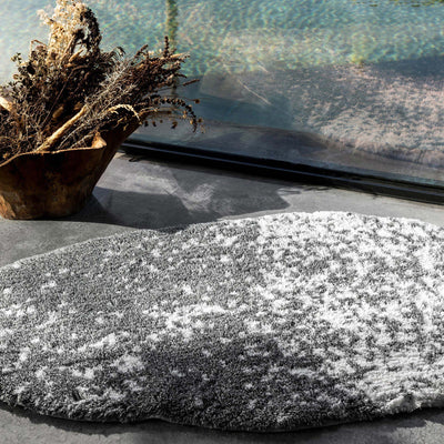 Stone bath rug in charcoal and white mottled texture, styled on stone floor near window.