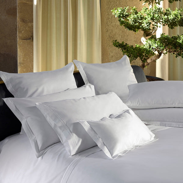 A neatly made bed with several white pillows and sheets, set near a window with beige curtains and a green potted plant in the background.