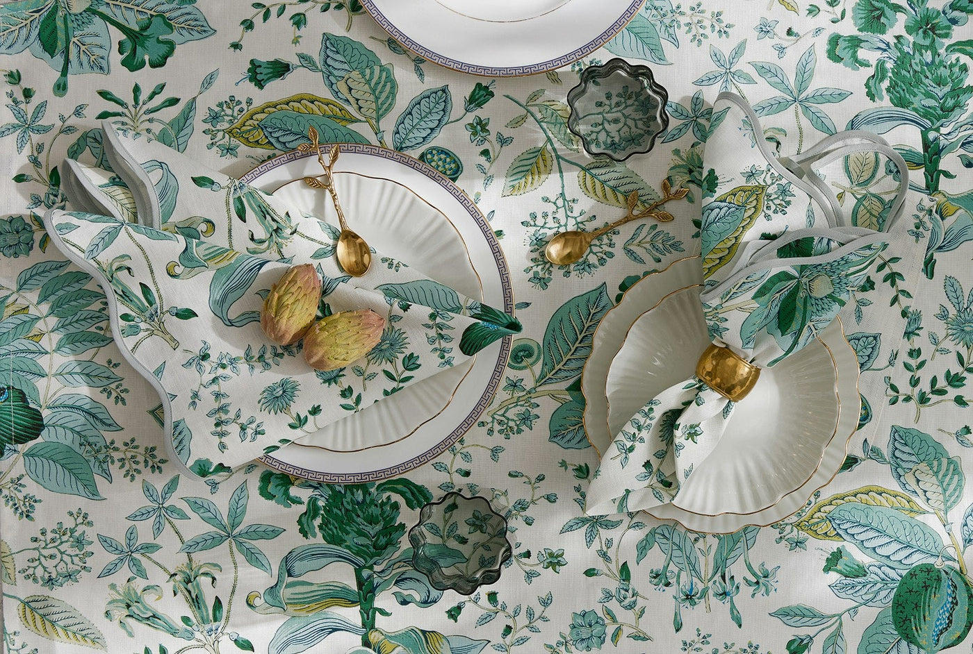 Overhead view of a table set with white plates, gold utensils, folded napkins, and a floral-patterned tablecloth in green and white.
