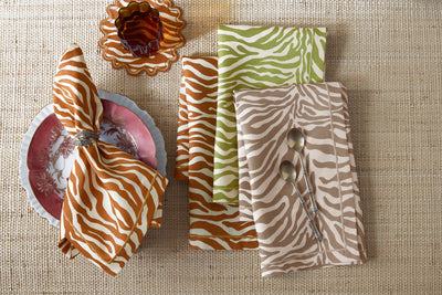 Table setting with a pink floral plate, silver napkin ring, glass, and zebra-patterned napkins in brown, green, and beige on a woven surface, with two spoons placed on the beige napkin.