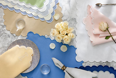 Table setting with scalloped-edge placemats in various colors, folded napkins, glassware, and a bouquet of cream roses on a marble surface.