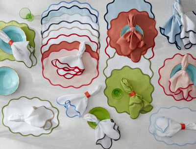 Overhead view of pastel plates with scalloped placemats and napkins in various colors, some tied with napkin rings, arranged neatly on a light surface.