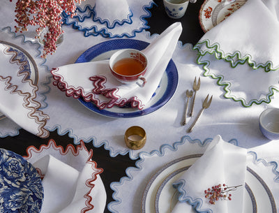 An overhead view of a table set with scalloped-edge white napkins, patterned plates, vintage cutlery, a cup of tea, and a small vase of red berries.