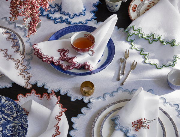 An overhead view of a table set with scalloped-edge white napkins, patterned plates, vintage cutlery, a cup of tea, and a small vase of red berries.