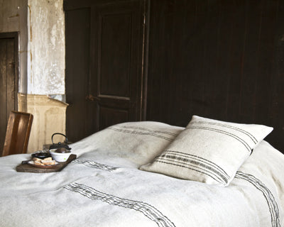 A neatly made bed with a white blanket featuring black stripes, a matching pillow, and a tray with tea and snacks placed on the blanket in a rustic room.