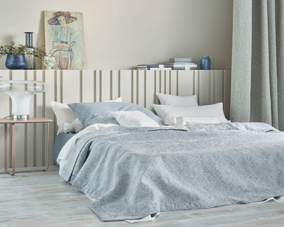 A neatly made bed with blue and white bedding, several pillows, a bedside lamp, vases, and a painting leaning on a ridged white headboard in a softly lit bedroom.