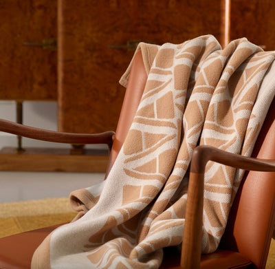 A beige and white patterned blanket draped over a tan leather armchair, with a wooden cabinet in the background.