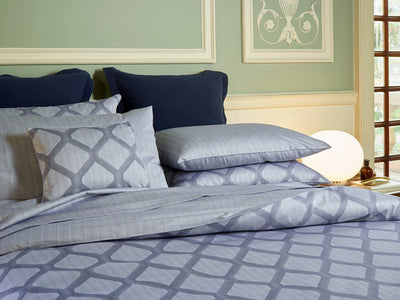 A neatly made bed with light blue and gray patterned bedding, navy pillows, and a round white lamp on a nightstand in a softly lit bedroom.
