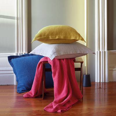 Three textured pillows—yellow, white, and blue—stacked on a wooden stool with a bright pink throw draped over it, set in a room with light walls and wood flooring.