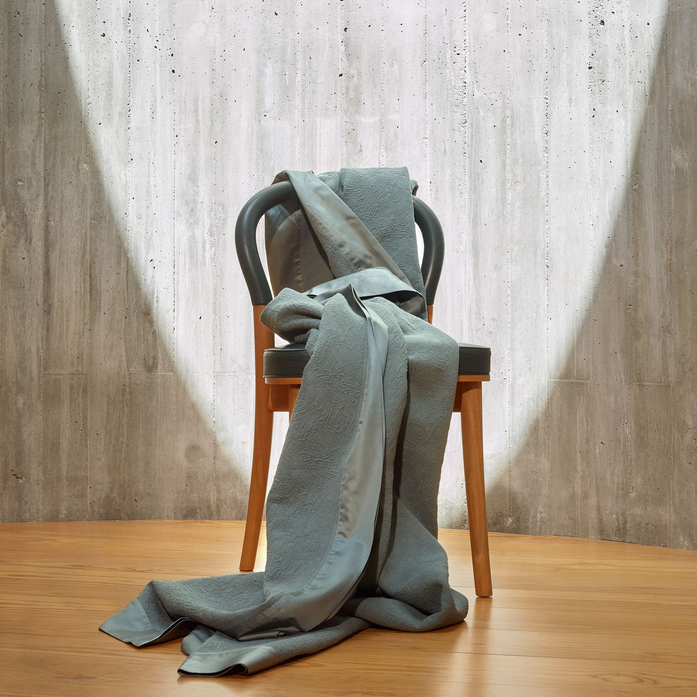 A light gray chair stands on a wooden floor with a matching gray blanket draped over and cascading onto the ground, set against a concrete wall with a circular spotlight.