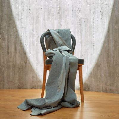 A light gray chair stands on a wooden floor with a matching gray blanket draped over and cascading onto the ground, set against a concrete wall with a circular spotlight.