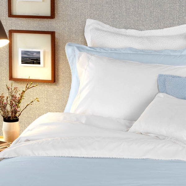 A neatly made bed with white and light blue bedding, pillows, a bedside lamp, a vase with flowers, and framed pictures on the wall.