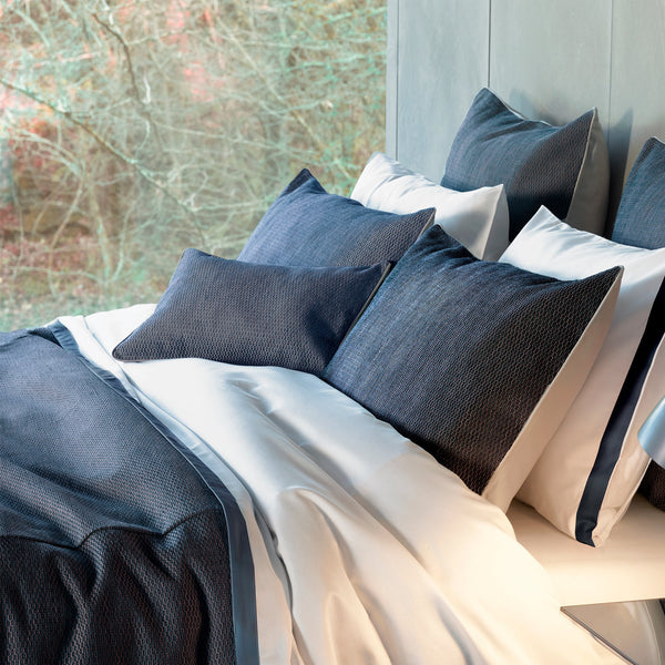 A neatly made bed with navy blue textured pillows and a matching comforter, set against a large window with a view of trees.