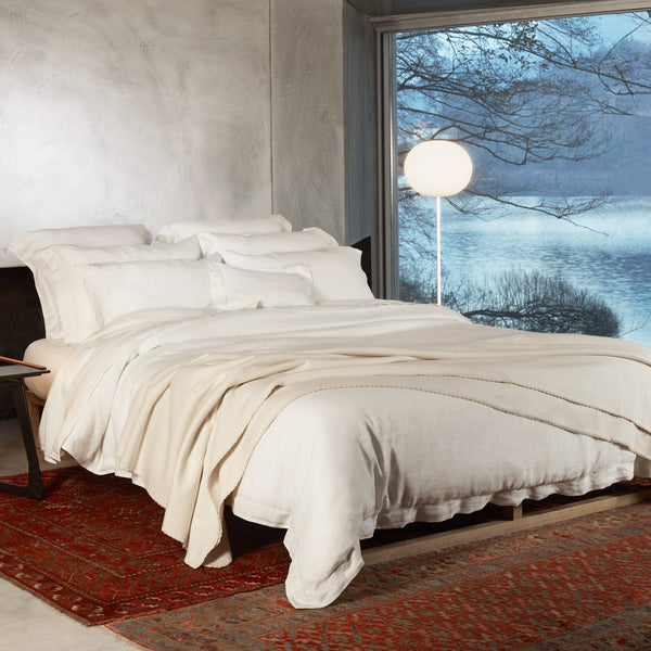 A neatly made bed with white bedding sits on a red patterned rug in a room with textured walls and a large window overlooking a lake and trees. A round floor lamp stands by the window.