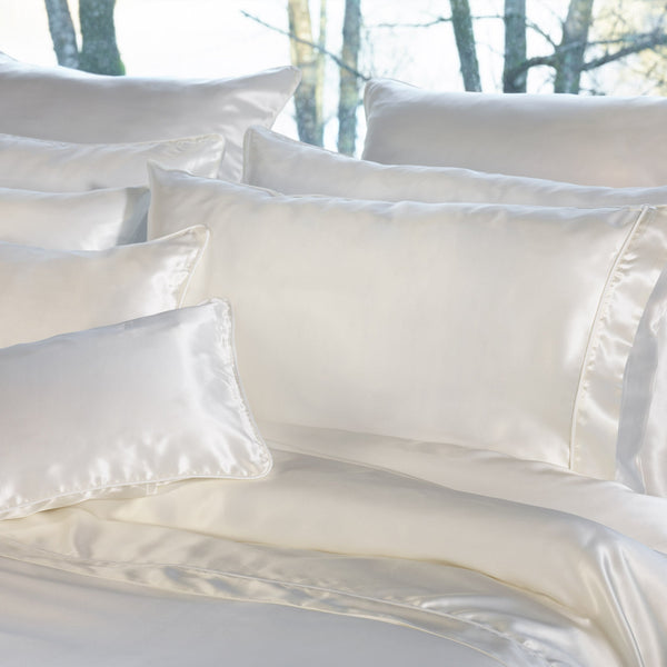 A neatly arranged bed features multiple white silk pillows and a matching silk duvet, with trees visible outside the window in the background.