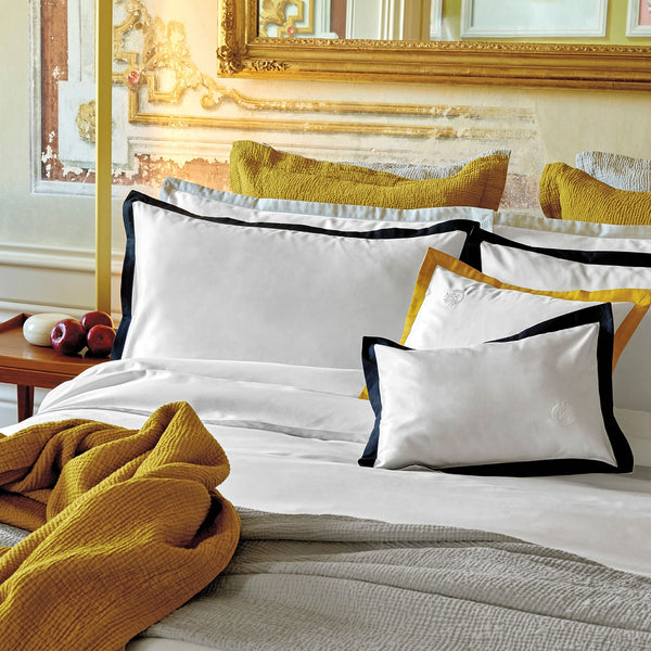 A neatly made bed with white sheets and pillows, some with black and yellow borders, a mustard yellow blanket, and a large ornate mirror on the wall above the headboard.