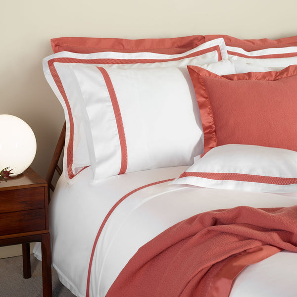 A neatly made bed with white bedding accented by coral trim and pillowcases, a matching coral blanket, and a round light on a wooden bedside table.