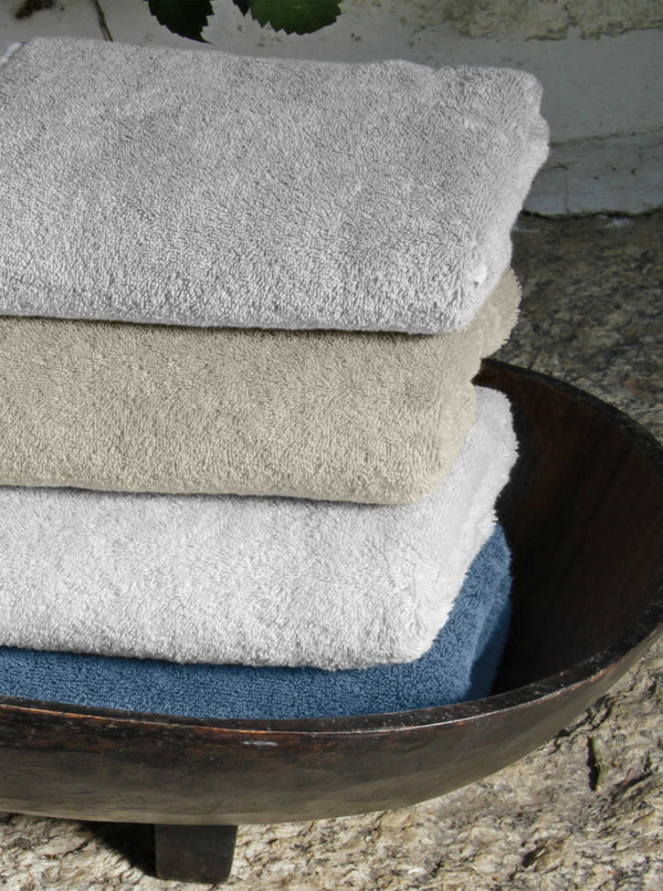 Four neatly folded towels in gray, beige, white, and blue are stacked in a dark wooden bowl on a stone surface.