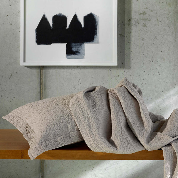 A beige textured pillow and blanket rest on a wooden surface with a monochrome abstract painting hanging on a concrete wall in the background.