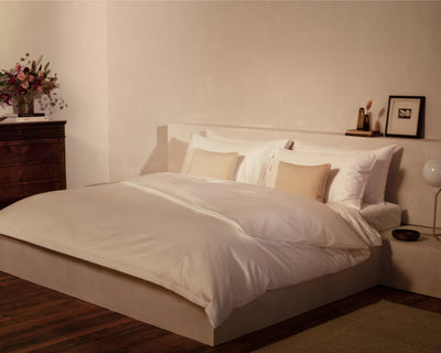 A neatly made bed with white bedding and beige pillows in a minimal bedroom. A wooden dresser with flowers and decor is beside the bed on a wooden floor.