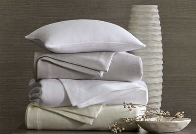Sferra Belem white and ivory folded sheet set stacked with pillow beside textured vase.