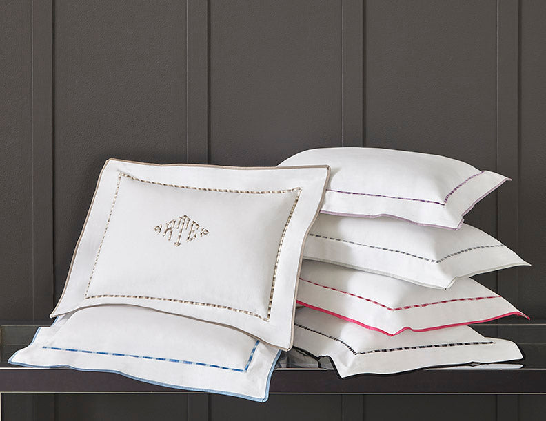 Sferra Calypso white pillowcases with colorful hemstitch trim stacked on dark bench.