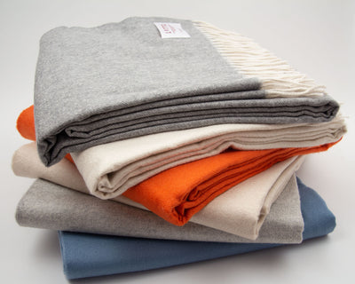 A neatly folded stack of wool blankets in grey, cream, orange, and blue, with one blanket showing white fringe on the edge.