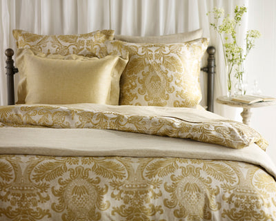 A neatly made bed with gold and beige damask-patterned bedding, two large pillows, and a small bedside table holding flowers and a book.