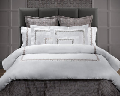 A neatly made bed with white bedding featuring a geometric border design, gray accent pillows, and a dark upholstered headboard against a paneled gray wall.