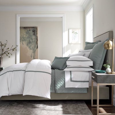 Sferra Fino white and sage cotton bedding collection styled on a bed in a neutral bedroom.