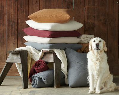 A golden retriever sits beside a rustic bench stacked with folded quilts and pillows in various neutral colors, set against a wooden wall.