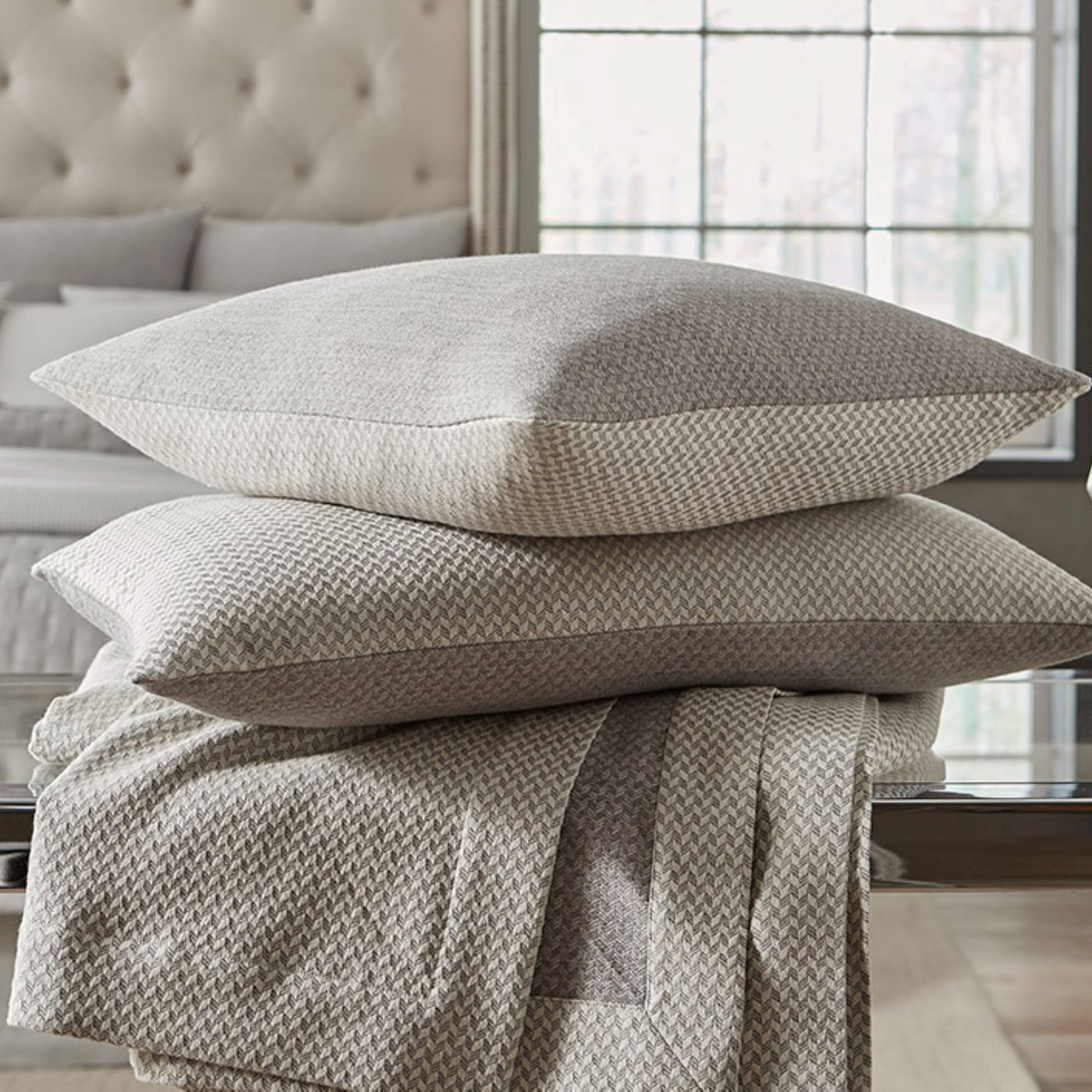 Sferra Geneva ivory herringbone cotton pillows and folded coverlet stacked on glass table.