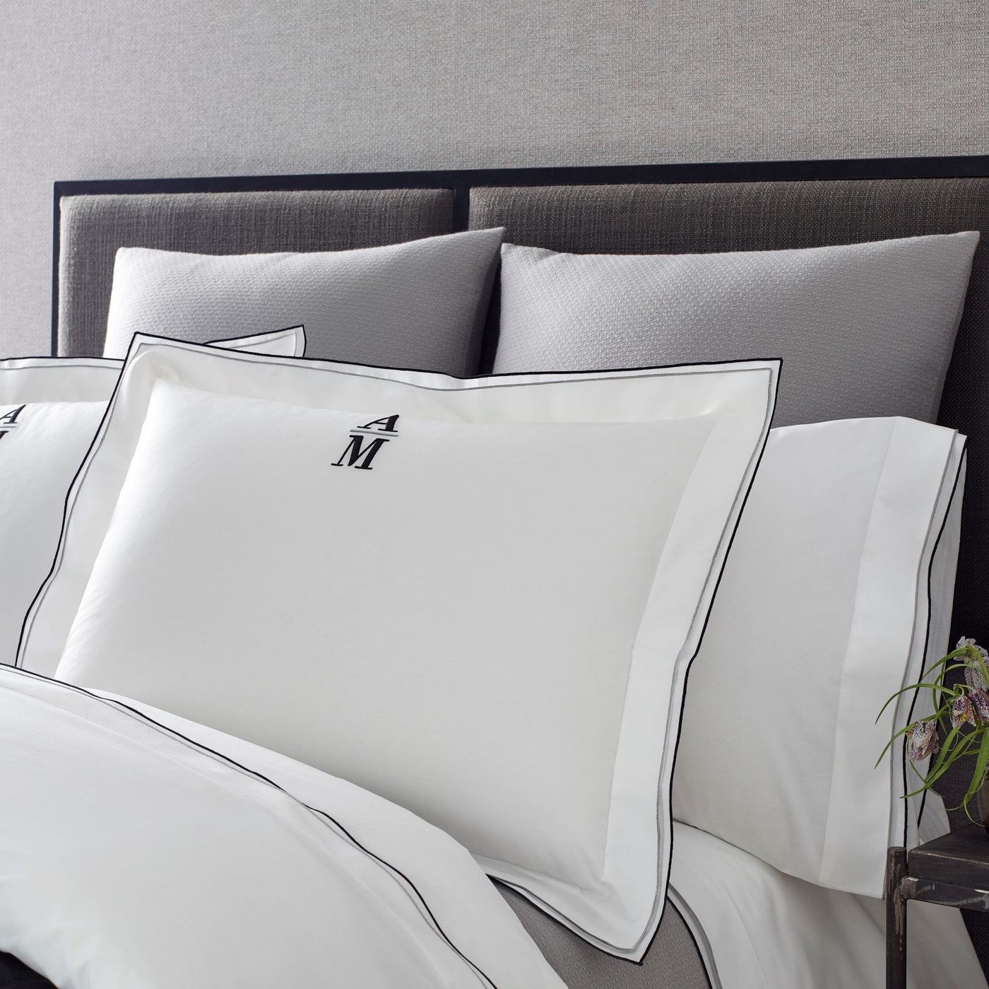 White cotton pillowcases with black double-stitch border and embroidered M monogram on made bed.