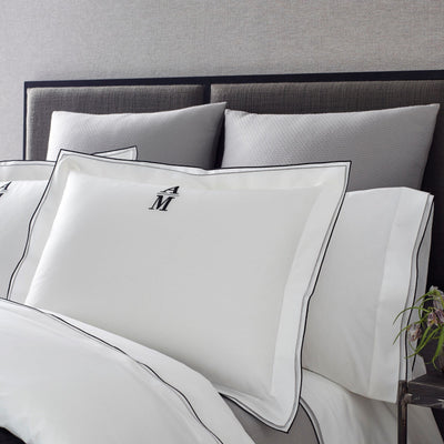 White cotton pillowcases with black double-stitch border and embroidered M monogram on made bed.