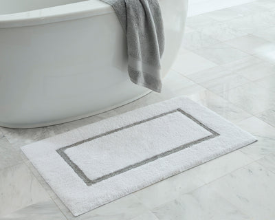 A white bath mat with a gray border is placed on a marble floor next to a white bathtub with a gray towel draped over the edge.