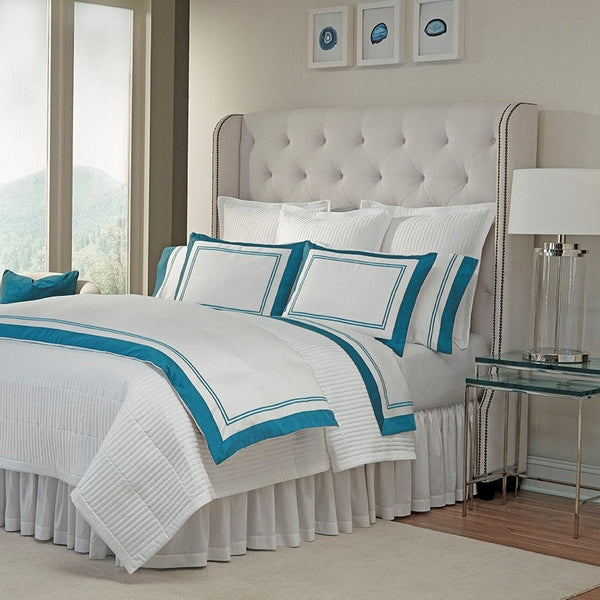 Home Treasures Marco white bedding collection with teal border on bed with tufted headboard.