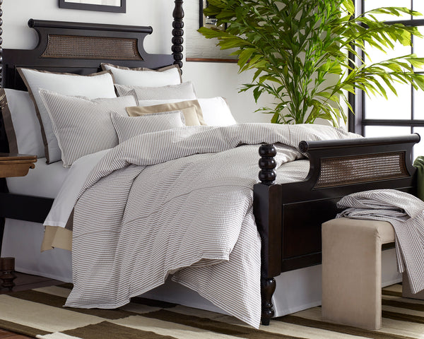 A neatly made bed with striped bedding and multiple pillows sits in a bright bedroom next to a large green plant and a beige ottoman.