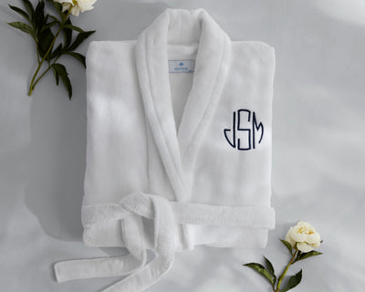 A white bathrobe with JSM monogram in navy blue is neatly folded on a marble surface, surrounded by white flowers and green leaves.