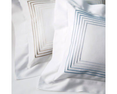 Two white pillow shams with embroidered square borders, one in taupe and one in light blue, placed side by side on a white surface.