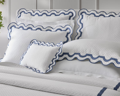 A neatly made bed with white quilted bedding and pillows featuring scalloped blue trim, set against a white paneled headboard.