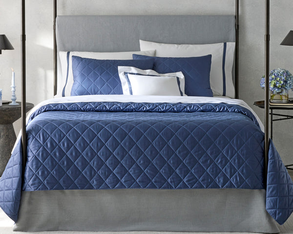 A neatly made bed with a grey upholstered headboard, navy blue quilted bedding, and several white and navy pillows. A side table holds a vase of flowers and a lit candle.