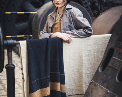 A person in work clothes stands with one arm resting on a metal rail, next to two draped woolen blankets, in an industrial setting with large machinery in the background.