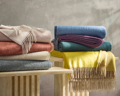 Stacks of folded wool blankets in various colors, including pink, blue, yellow, beige, and gray, are arranged on two wooden tables against a neutral background.
