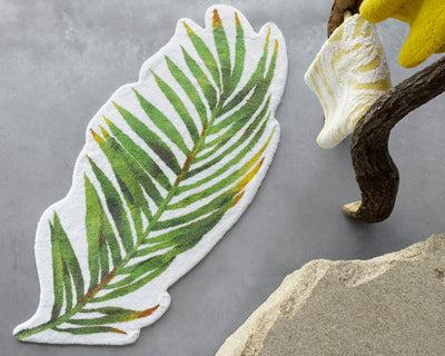A leaf-shaped rug with green and yellow detailing lies on a concrete floor next to a tree branch and a textured rock.