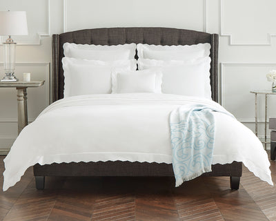 A neatly made bed with white scalloped bedding and pillows, a dark upholstered headboard, a light blue patterned throw, and a bedside table with a lamp and candle.